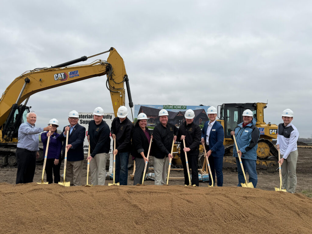 Participants wearing hard hats hold shovels in dirt during an outdoor groundbreaking ceremony, with construction equipment behind them.